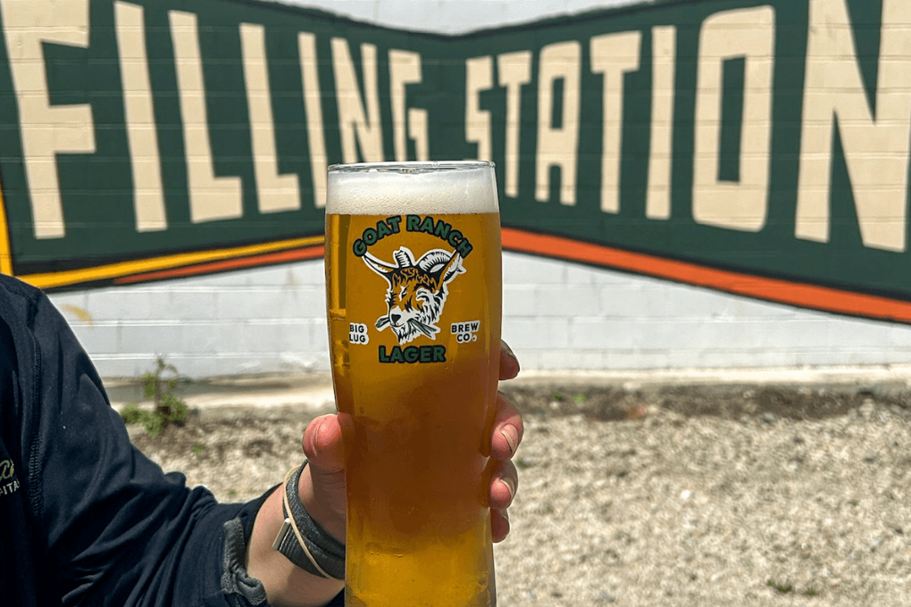 a hand holding up a tall frosty 21oz pour of Goat Ranch Lager in front of the building mural that reads 